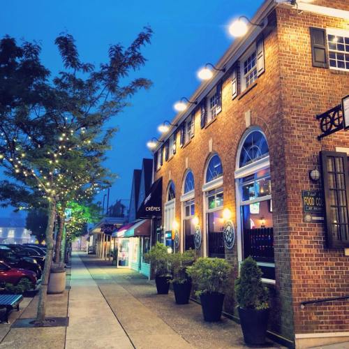 A charming street scene at dusk, featuring brick buildings, string lights, and outdoor dining areas.