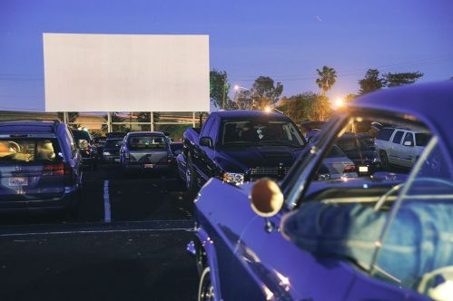 A drive-in movie scene at dusk, featuring parked cars and a blank screen in the background.
