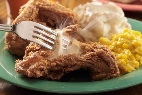 A plate of crispy fried chicken with mashed potatoes and corn, steam rising from the chicken as a fork pierces it.