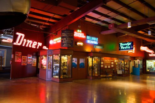 A vibrant indoor scene featuring neon signs for a diner and various businesses, with a nostalgic atmosphere.