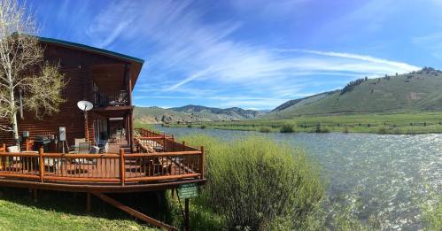 A scenic view of a wooden cabin by a river, surrounded by green hills and a blue sky with wispy clouds.