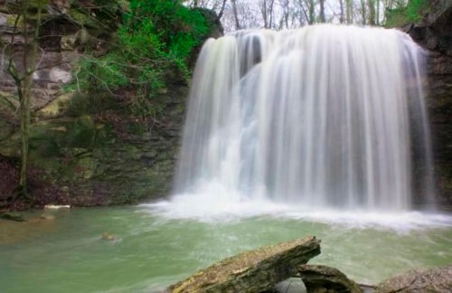 A serene waterfall cascades over rocky cliffs into a calm, green pool surrounded by lush trees.
