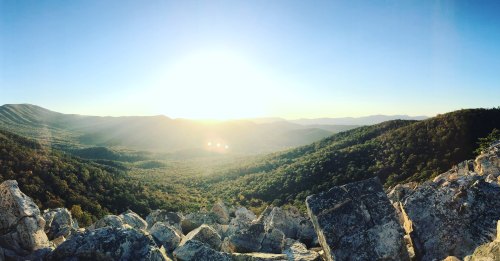 A panoramic view of mountains and valleys at sunrise, with sunlight illuminating the landscape and rocky foreground.