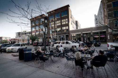 Outdoor dining scene with people at tables, modern buildings, and parked cars on a sunny day.