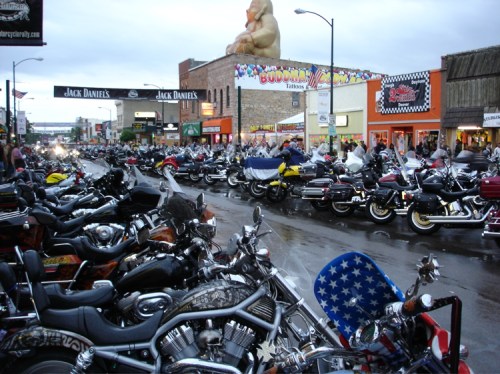 A bustling street filled with rows of parked motorcycles, with shops and a large statue in the background.