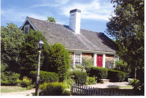 A charming house with a red door, surrounded by lush greenery and a white picket fence under a clear blue sky.
