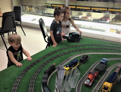 Three children play with a model train set on a green table, observing trains on tracks in a display area.