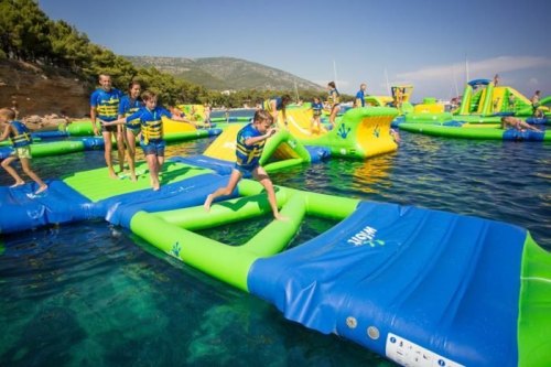 Children and adults play on colorful inflatable water structures in a sunny, blue water setting.