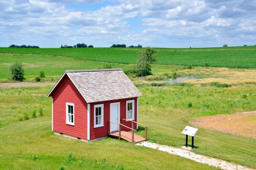 A small red house with a porch sits in a green field under a cloudy sky, surrounded by lush farmland.