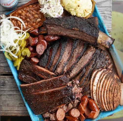 A colorful platter of smoked meats, including brisket, ribs, sausage, and sides of beans, coleslaw, and pickles.
