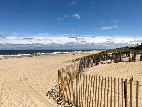 A sandy beach with gentle waves, a blue sky, and a wooden fence along the shore, creating a serene coastal scene.