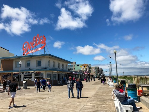 A bustling boardwalk scene with shops, people walking, and a bright blue sky with clouds.
