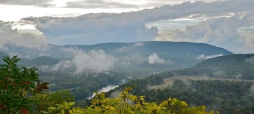 A scenic view of rolling hills and a river, with clouds and mist hovering over the landscape.