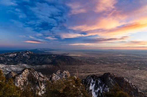A panoramic view of snow-capped mountains under a colorful sunset sky, overlooking a vast valley below.