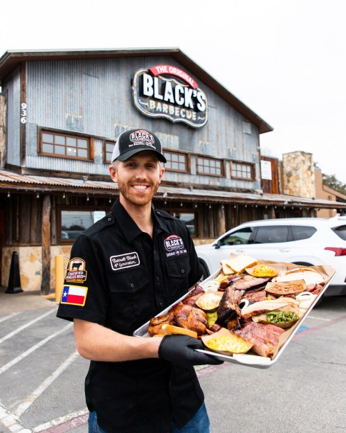 A smiling man in a black shirt holds a large platter of barbecue outside a rustic restaurant.