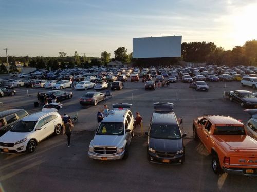 A busy drive-in movie theater parking lot at sunset, with cars parked and people gathering outside.