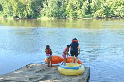 Children playing by a river, preparing to use colorful inflatable tubes on a wooden dock surrounded by greenery.