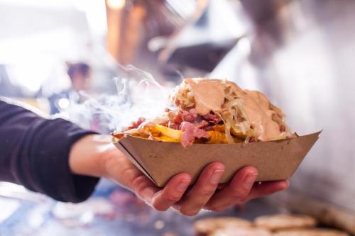 A hand holding a bowl of loaded nachos topped with sauce, cheese, and toppings, with steam rising in the background.