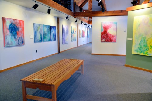 Art gallery interior featuring colorful abstract paintings on the walls and a wooden bench in the center.