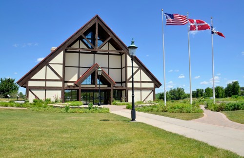 A large, modern building with a triangular roof, surrounded by green grass and flower beds, featuring three flags out front.