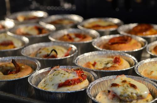 Baked mini pizzas in aluminum cups, topped with cheese, pepperoni, and herbs, arranged in a row.