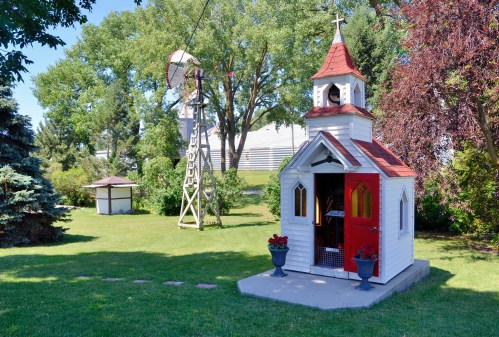 A small white chapel with a red door, surrounded by greenery and a windmill in the background.
