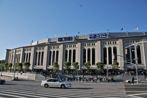 Exterior view of a large stadium with banners, trees, and a line of people outside on a sunny day.