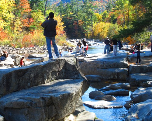 A scenic river with people enjoying the outdoors, surrounded by colorful autumn foliage and rocky terrain.