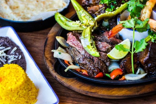 A sizzling platter of grilled meats, peppers, and onions, served with rice, beans, and a side of tortillas.