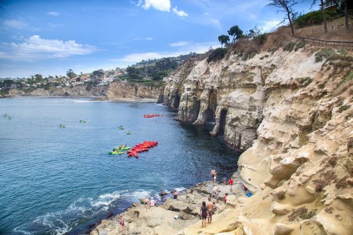 Kayakers in bright boats near rocky cliffs, with people exploring the shore and a scenic coastal backdrop.