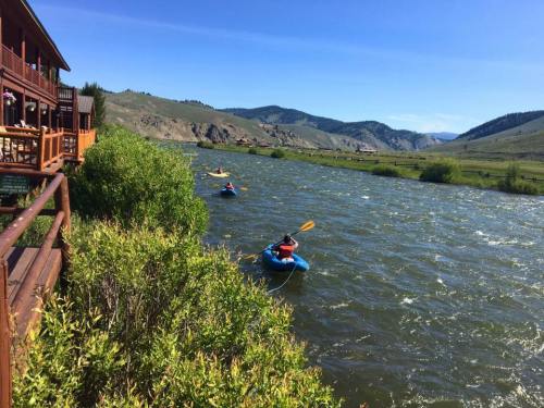 Two kayakers paddle on a river surrounded by green foliage and mountains under a clear blue sky.