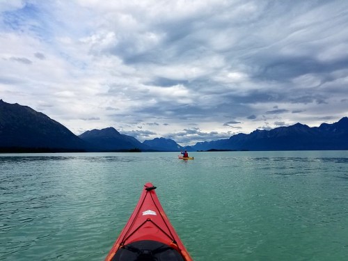 A red kayak on calm water, with mountains and a cloudy sky in the background. Another kayaker is visible in the distance.