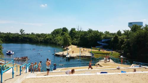 A sunny beach scene with people swimming and relaxing by a lake, surrounded by trees and a water slide in the background.