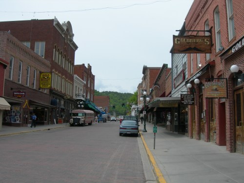 A historic street lined with brick buildings, shops, and a vintage bus parked on the side, under a cloudy sky.