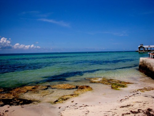 A serene beach scene with clear blue water, rocky shore, and a bright sky dotted with clouds.