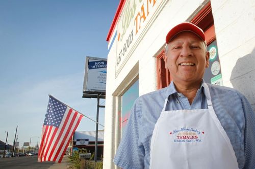 A smiling man in an apron stands outside a tamale shop, with an American flag waving in the background.