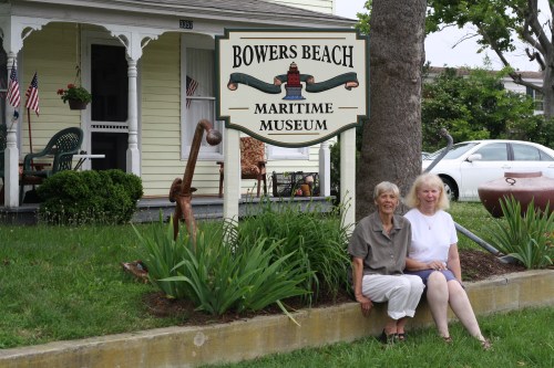 Two women sit on the grass in front of the Bowers Beach Maritime Museum sign, with a house and greenery in the background.