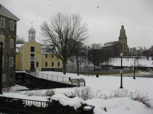 A snowy landscape featuring a yellow building, a church, and bare trees by a frozen body of water.