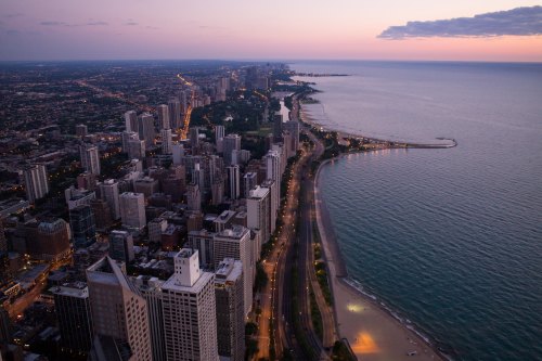 Aerial view of a city skyline at dusk, with buildings along the shore and a calm lake reflecting the evening light.