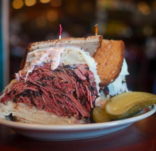 A stacked sandwich with layers of pastrami, sauerkraut, and pickles on a plate, garnished with toothpicks.