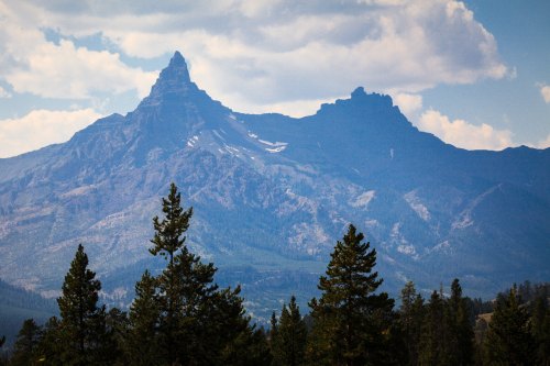 A majestic mountain range with a prominent peak, surrounded by lush green trees and a blue sky with clouds.