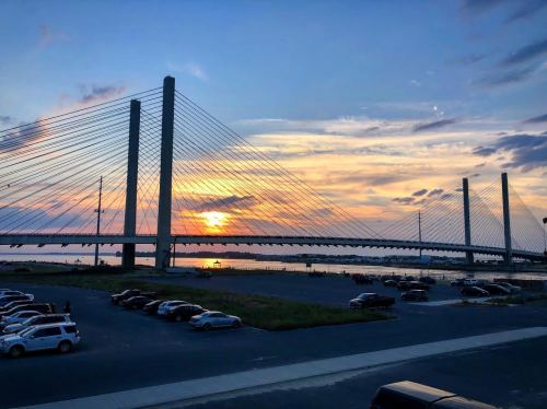 A sunset view behind a cable-stayed bridge, with cars parked in the foreground and clouds in the sky.