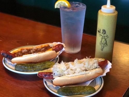 Two hot dogs with toppings, pickles on the side, a glass of water, and a bottle of mustard on a wooden table.