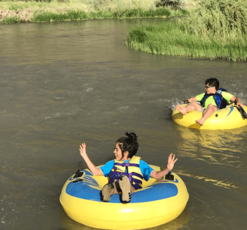 Two children in yellow inner tubes float down a river, surrounded by greenery and enjoying a sunny day.