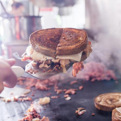 A hand holding a spatula with a grilled sandwich, steam rising from a hot cooking surface in the background.