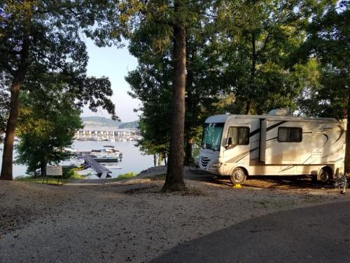An RV parked among trees with a view of a marina and calm water in the background.