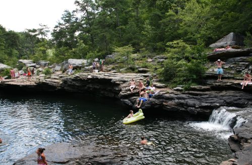 A natural swimming area with people relaxing on rocks, swimming, and enjoying the water surrounded by lush greenery.