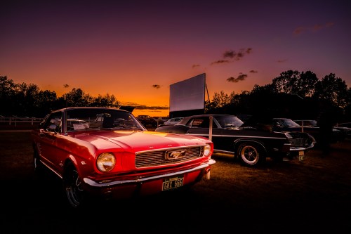 A red classic Mustang and a black vintage car parked at sunset, with a drive-in movie screen in the background.
