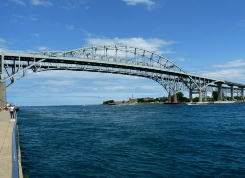 A large steel bridge spans over a blue body of water under a partly cloudy sky, with people walking along the shore.
