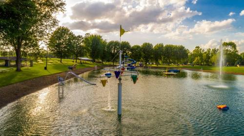 A scenic park with a pond, water slide, and colorful water features under a partly cloudy sky.